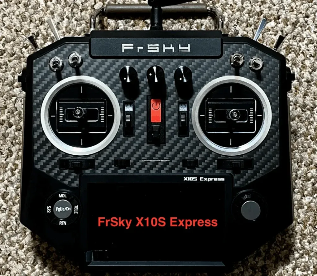 A sophisticated FrSky X10S Express remote control rests on a carpeted surface, its black facade with carbon fiber patterns exuding a sleek, professional aura. The dual control sticks, framed by silver bezels, await the pilot’s command, while the top switches and screen displaying ‘FrSky X10S Express’ promise precise control over distant flights. Below, the MDL, PgUp/PgDn, and RTN buttons hint at the complex functionality housed within this compact device.